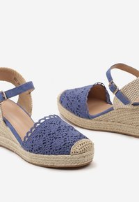 Sandales en crochet bleu à talon compensé avec un bout fermé, plateforme en jute et bride de cheville. Présente un motif texturé sur le dessus.