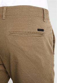 Bruine katoenen broek met een achterzak met een verborgen naad en een klein zwart label bevestigd aan de tailleband.