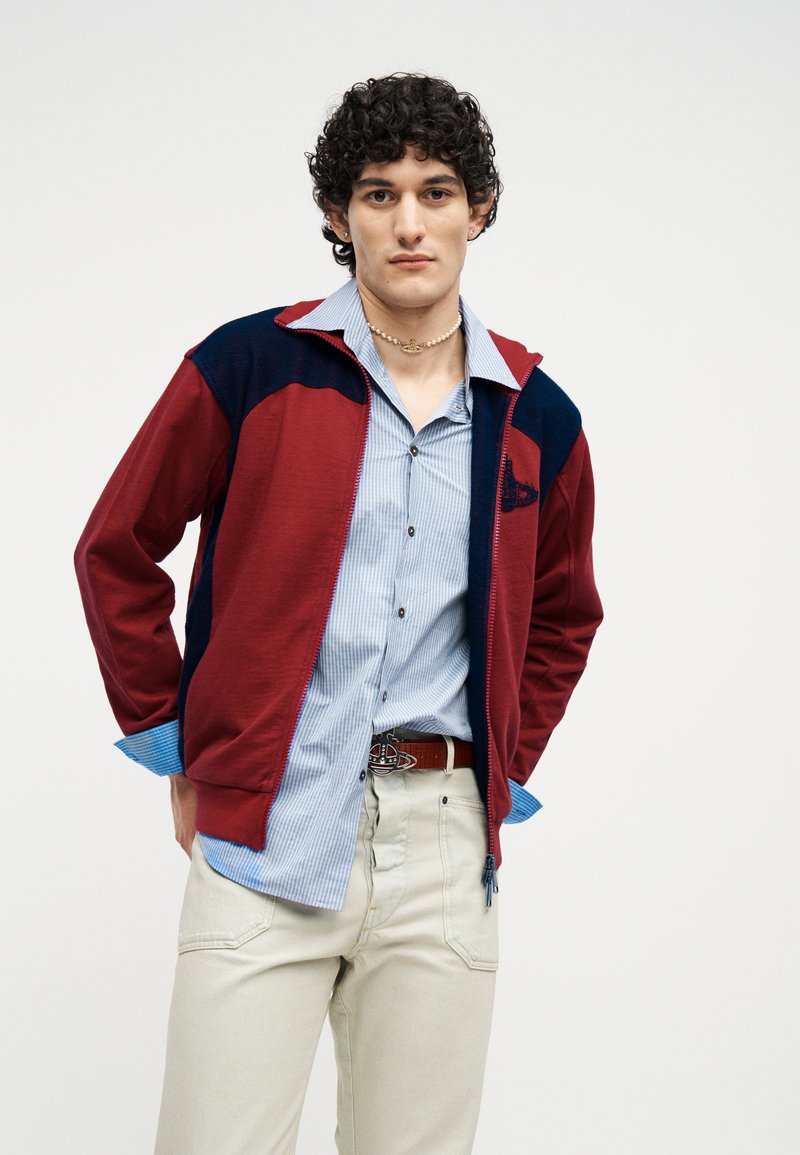 Young man with curly hair wearing a red and navy jacket over a blue striped shirt and beige pants, standing against a plain light background.