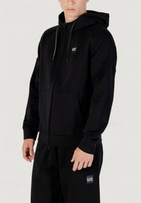Schwarze Kapuzenjacke mit einer Kapuze, aus glattem Stoff gefertigt, verfügt über Fronttaschen, gerippte Bündchen und ein kleines Logopatch auf der Brust.