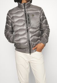 Giacca a vento grigia con colletto alto, design trapuntato, zip frontale e tasca con zip nera. Polsini e orlo a costine.