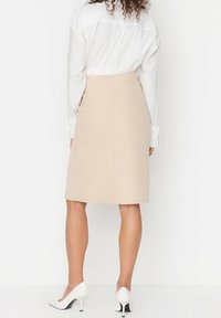 Jupe crayon beige jusqu'au genou avec une texture lisse et une fermeture éclair discrète à l'arrière, associée à une chemise blanche et des chaussures à talons blancs.