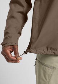 Giacca impermeabile marrone con vita regolabile, texture liscia e polsini regolabili. Zip con impugnature manuali all'orlo.