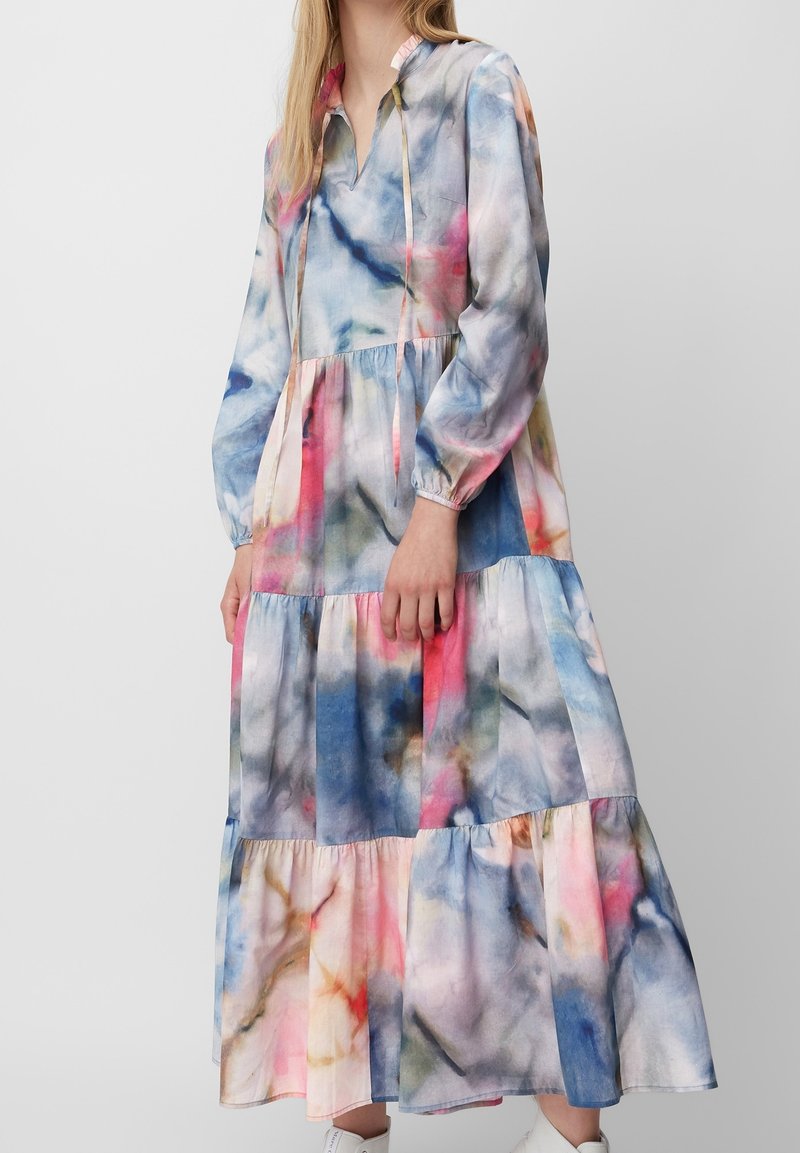 Femme portant une robe maxi à manches longues et à volants avec un motif aquarelle multicolore et une coupe ample, debout devant un fond uni.
