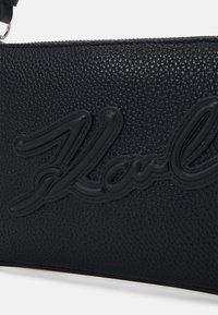 Svart texturerad läderväska med en framträdande präglad varumärkesnamn i en matchande mörk ton. Slät dragkedja med en elegant design.