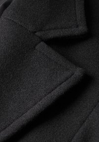 Primer plano de un abrigo de lana negro que destaca su suave textura, solapas afiladas, costuras limpias y bordes precisos. El material parece grueso y cálido.