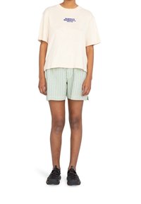 Crème t-shirt met korte mouwen en blauwe opdruk "ELEMENT Skateboard Co.", gecombineerd met groene gestreepte shorts en zwarte sportschoenen.