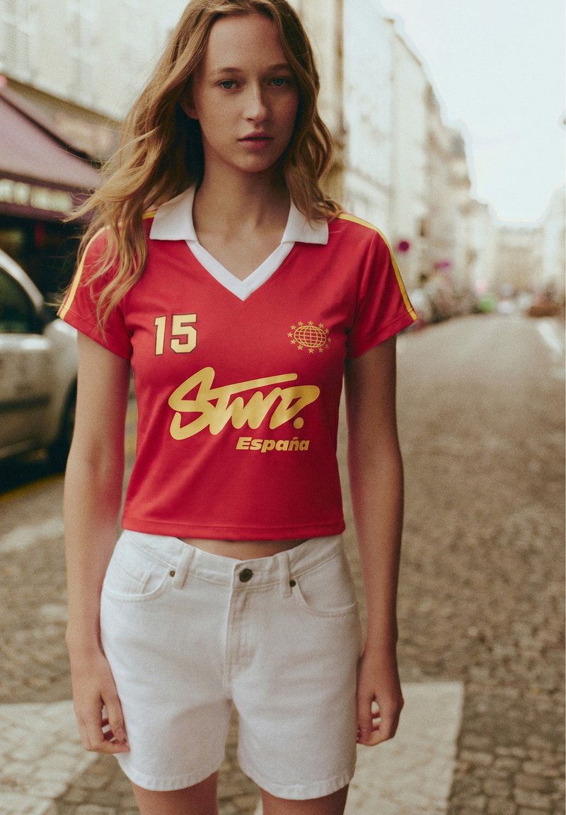 Jeune femme aux longs cheveux blonds porte un haut de sport rouge court avec « España » et un short blanc dans une rue de la ville.