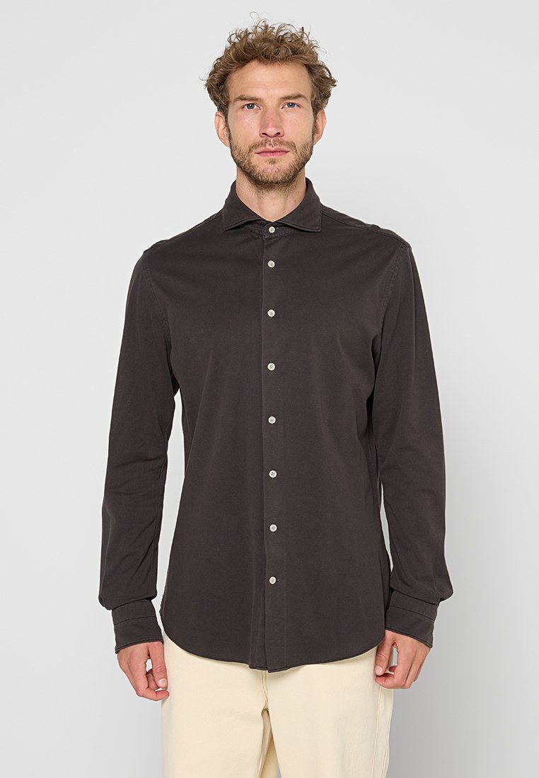 Man met krullend haar, gekleed in een zwarte knoop-overhemd met lange mouwen en beige broek, staande tegen een lichte egale achtergrond.