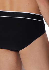 Zwarte zwemslips met een witte elastische tailleband. De stof is glad en heeft een strakke pasvorm, met een minimalistisch ontwerp zonder extra patronen.
