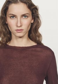 Burgunderfarbener Langarmtop mit weiter Ausschnitt, aus leichtem, leicht durchscheinendem Stoff, mit weicher Textur und ungefinishte Kanten.