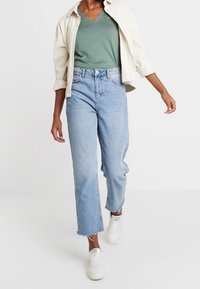 Ljusblå denimjeans med rak passform och fransad nederkant, kombinerat med en grön t-shirt med V-hals och en off-white knapp-up skjorta.