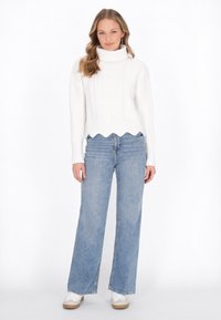Weißer gestrickter Pullover mit Zopfstrickmuster und Rollkragen, kombiniert mit hellblauen weiten Jeans und weißen Sneakers. Scallop-Saum des Pullovers.