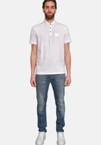 Weißes Poloshirt mit Kragen, drei Knöpfen und kleinem Logo auf der linken Brust. Kombiniert mit mittelblauen Jeans und weißen Sneakers.