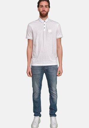 Weißes Poloshirt mit Kragen, drei Knöpfen und kleinem Logo auf der linken Brust. Kombiniert mit mittelblauen Jeans und weißen Sneakers.