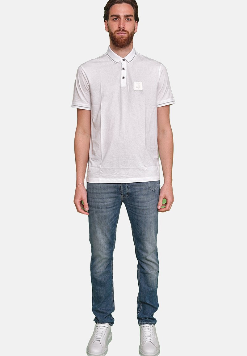 Weißes Poloshirt mit Kragen, drei Knöpfen und kleinem Logo auf der linken Brust. Kombiniert mit mittelblauen Jeans und weißen Sneakers.