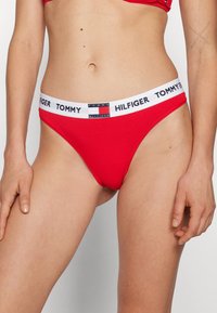 Un string rouge en coton, doté d'une large ceinture blanche avec le logo "TOMMY HILFIGER" et un design minimaliste.