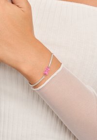 Silbernes Armband mit klaren Strasssteinen und einem rosa Gummibärchen-Anhänger, getragen an einer Hand mit einem weißen, strukturierten Ärmel.