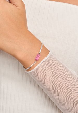 Silbernes Armband mit klaren Strasssteinen und einem rosa Gummibärchen-Anhänger, getragen an einer Hand mit einem weißen, strukturierten Ärmel.