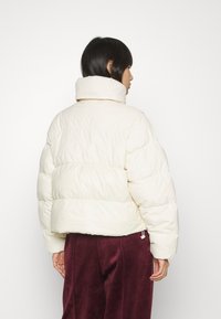 Doudoune off-white, avec un col montant, des sections matelassées et un ourlet ajustable. Associée à un pantalon en tissu texturé de couleur sombre.