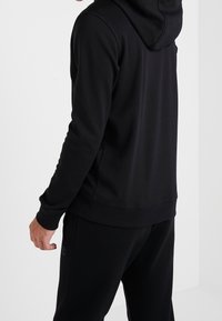 Sudadera con capucha negra de textura suave, con puños ajustados y cinturilla. Diseño sencillo, sin logotipos ni patrones visibles, combinada con pantalones a juego.