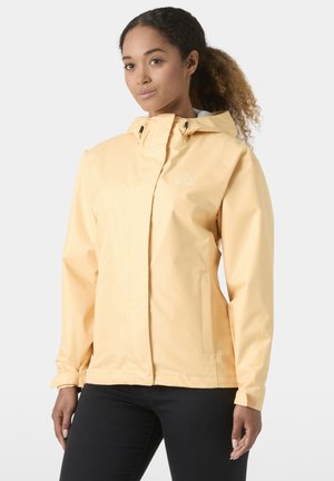 Woman with curly hair wearing a light yellow hooded rain jacket and black pants, standing against a plain light background.