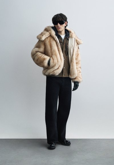 Παλτό από τεχνητή γούνα σε ανοιχτό καφέ χρώμα με κουκούλα, φορεμένο πάνω από σκούρο πουκάμισο και πουλόβερ με στρογγυλή λαιμόκοψη. Μαύρο παντελόνι φαρδύ και γάντια ολοκληρώνουν την εμφάνιση.