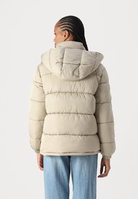 Beige dunjakke med hette, med quiltede seksjoner og elastiske mansjetter. Mykt materiale og avslappet passform, sammen med lyseblå jeans.