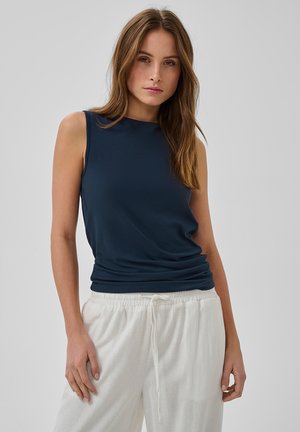 Femme aux longs cheveux bruns portant un haut sans manches bleu marine et un pantalon blanc avec cordon de serrage, debout devant un fond gris uni.