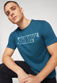 Türkiser T-Shirt mit kurzen Ärmeln aus Baumwolle, ausgestattet mit einem weißen Grafikdruck auf der Brust. Das Modell kniet und hält die Hände auf den Beinen.