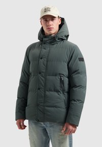 Gevoerde groene jas met een capuchon, glanzende textuur, knoop- en ritsafsluitingen, en twee zijzakken. gedragen over lichtblauwe versleten jeans.