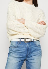 Pull en tricot blanc avec un motif en câble, porté sur un jean en denim bleu, arborant une ceinture blanche avec une boucle en argent décorative.