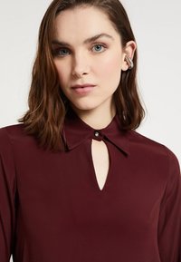 Bordowa bluzka z małym detalem w kształcie otworu przy szyi, kołnierzykiem zapinanym na guzik oraz długimi rękawami wykonanymi z gładkiego materiału.