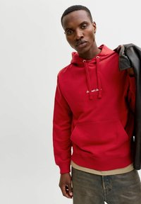 Sweatshirt à capuche rouge avec cordons, poche avant et texte brodé. Porté sur une chemise beige avec un pantalon en denim et une veste noire.