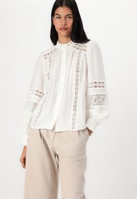 White blouse with a high neckline, intricate lace trim, button front, and puffed sleeves. Paired with light beige trousers.
