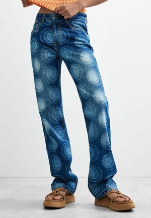 Persoon die een blauwe spijkerbroek met ronde patronen, een oranje gestreepte crop top en bruine platformsandalen draagt, staand op een grijze vloer.