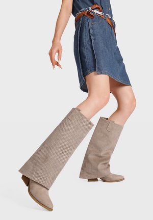 Mujer que lleva una falda vaquera y botas perforadas marrón claro hasta la rodilla, dando un paso adelante sobre un fondo liso.