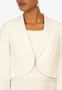 Hvid cropped blazer med spidse klapper, glat tekstur og enkel lukning. Bærer den over en hvid top med kvadratisk udskæring.