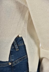 Maglione lavorato a maglia color crema con motivi testurizzati, taglio laterale e dettaglio con perla. Abbinato a pantaloni di denim blu.