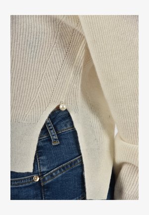 Pleťový pulóver z pleteniny krémovej farby s textúrovanými vzormi, bočným výrezom a perlovým doplnkom. Kombinovaný s modrými džínsovými nohavicami.