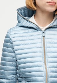 Γυναίκα που φοράει ανοιχτό μπλε μπουφάν με κουκούλα και μπεζ φερμουάρ, ορατή από τον λαιμό έως το μέσο του κορμού, μπροστά σε απλό φόντο.