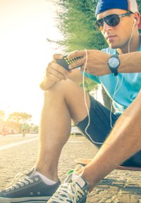 Young man wearing black sunglasses and a blue shirt, holding a yellow and black device, with earphones, seated on a sidewalk.
