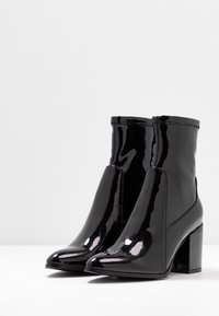 Bottes cheville en cuir verni noir avec bout pointu, talon haut et fermeture éclair latérale, présentant une texture lisse et un design minimaliste.
