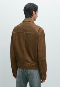 Brun mockasinjacka med krage, sidofickor och knäppte ärmar. Avslutad med en ren design och en mjuk textur. Bärs med ljusgrå byxor.