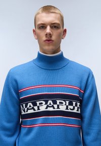 Pullover blu a maglia con colletto alto, caratterizzato da righe orizzontali in rosso, blu e nero, e una banda con logo bianco che attraversa il petto.