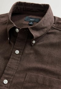 Brun skjorta med knappar gjord av texturerad tyg, med spetsig krage, vita knappar och en bröstficka. Varumärkesetikett synlig inuti kragen.