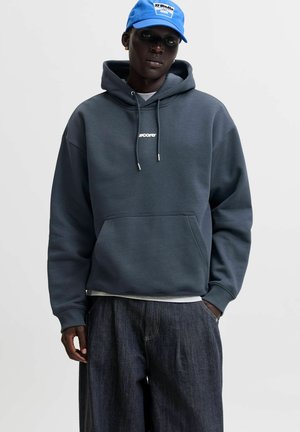Homme portant un sweat à capuche gris foncé avec poche avant, un jean sombre ample et une casquette bleu vif, debout devant un fond clair uni.