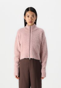 Suéter corto de color rosa claro, hecho de un material suave, que cuenta con un cuello alto y una cremallera en el frente, con puños y dobladillo acanalados. Combinado con pantalones marrones.