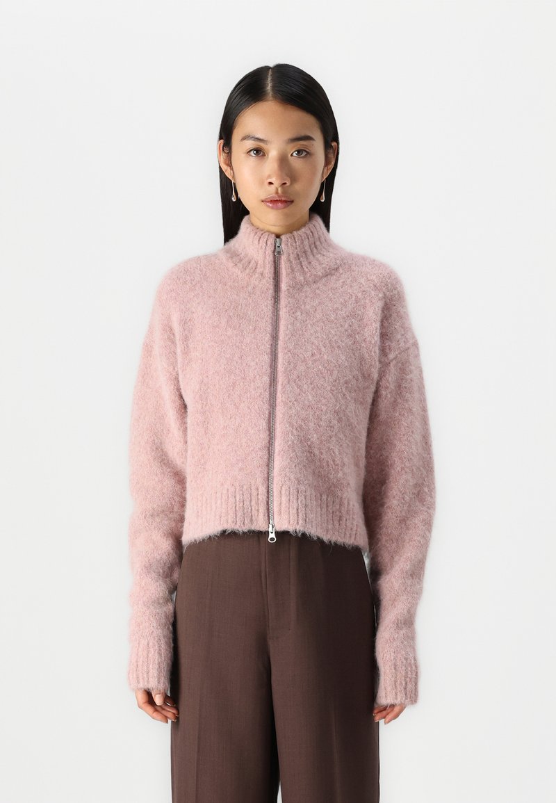 Suéter corto de color rosa claro, hecho de un material suave, que cuenta con un cuello alto y una cremallera en el frente, con puños y dobladillo acanalados. Combinado con pantalones marrones.