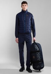 Gilet imbottito blu navy con trapuntatura orizzontale, chiusura con zip e patch con logo, abbinato a pantaloni blu navy e una grande borsa da viaggio. Scarpe nere.
